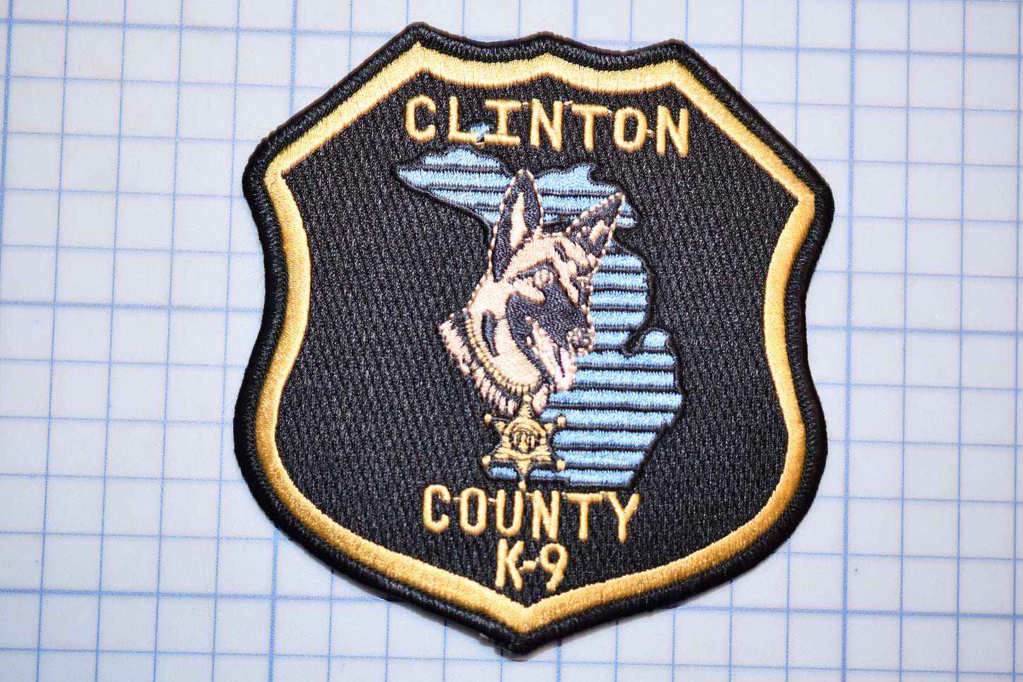 A black and gold badge with a dog and the text "CLINTON COUNTY K-9" on it.
