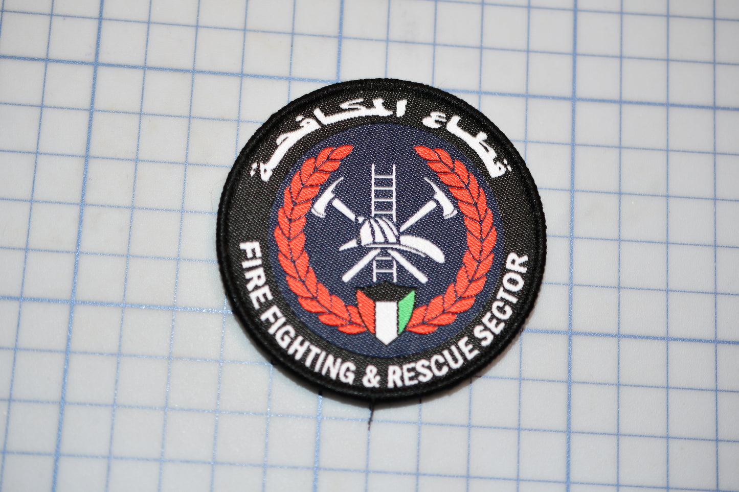 a circular patch with a logo, surrounded by a wreath and text, placed on a grid background.