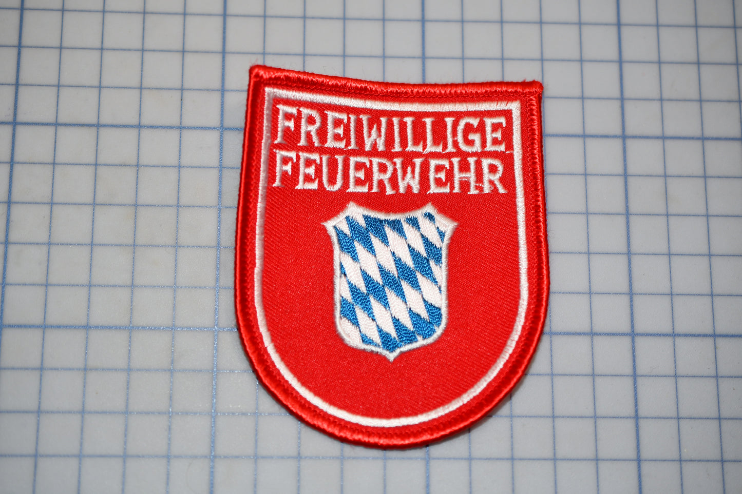 a red badge with the text "Freiwillige Feuerwehr" on it, which is a German phrase that translates to "Free will" in English. The badge is displayed on a grid background.