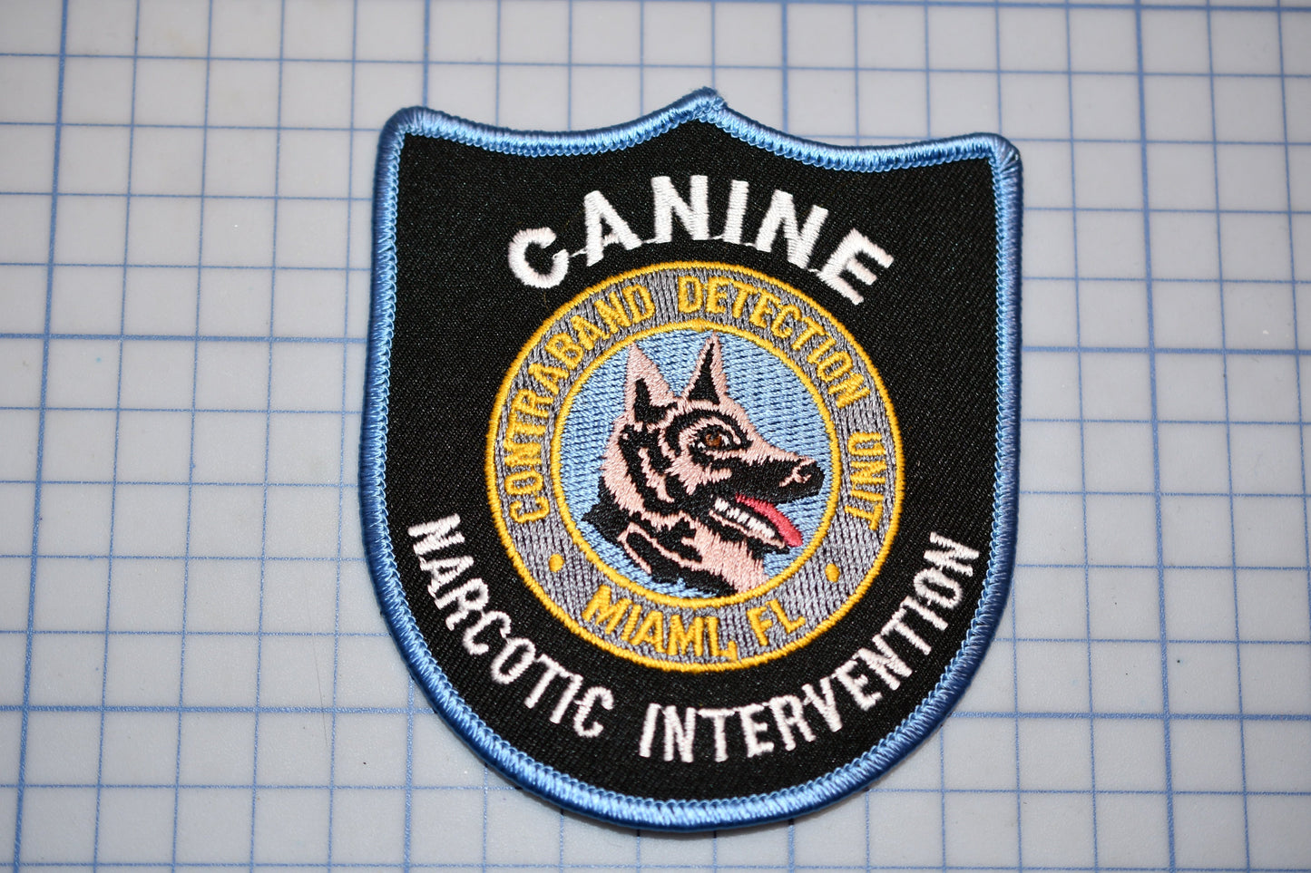 A black and blue patch with the text "CANINE DETECTION UNIT" and "NARCOTIC INTERVENTION" printed on it, along with a graphic of a dog's head.