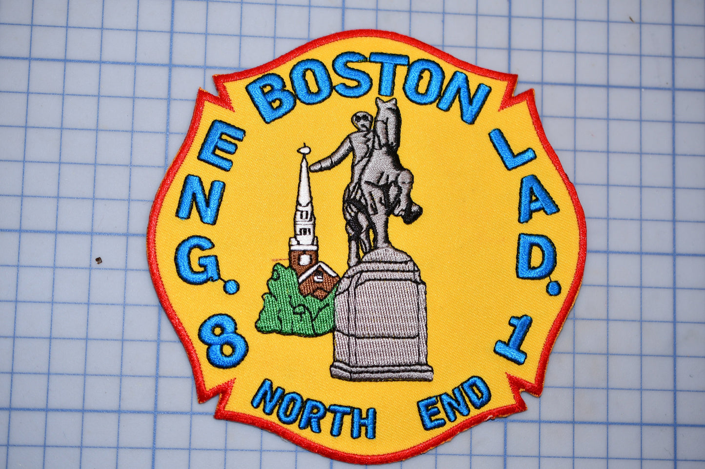 a fire department patch with a yellow background and a red border. The patch displays a statue of a man on a horse, a church steeple, and a building. The text on the patch reads "BOSTON LAD. 1 NORTH END".