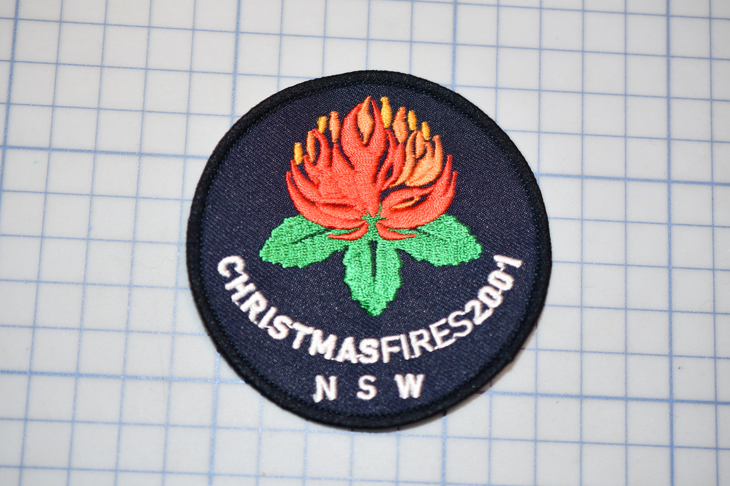 A circular patch with a red flower and green leaves, along with the text "CHRISTMASFIRES2001 N SW" printed on it.