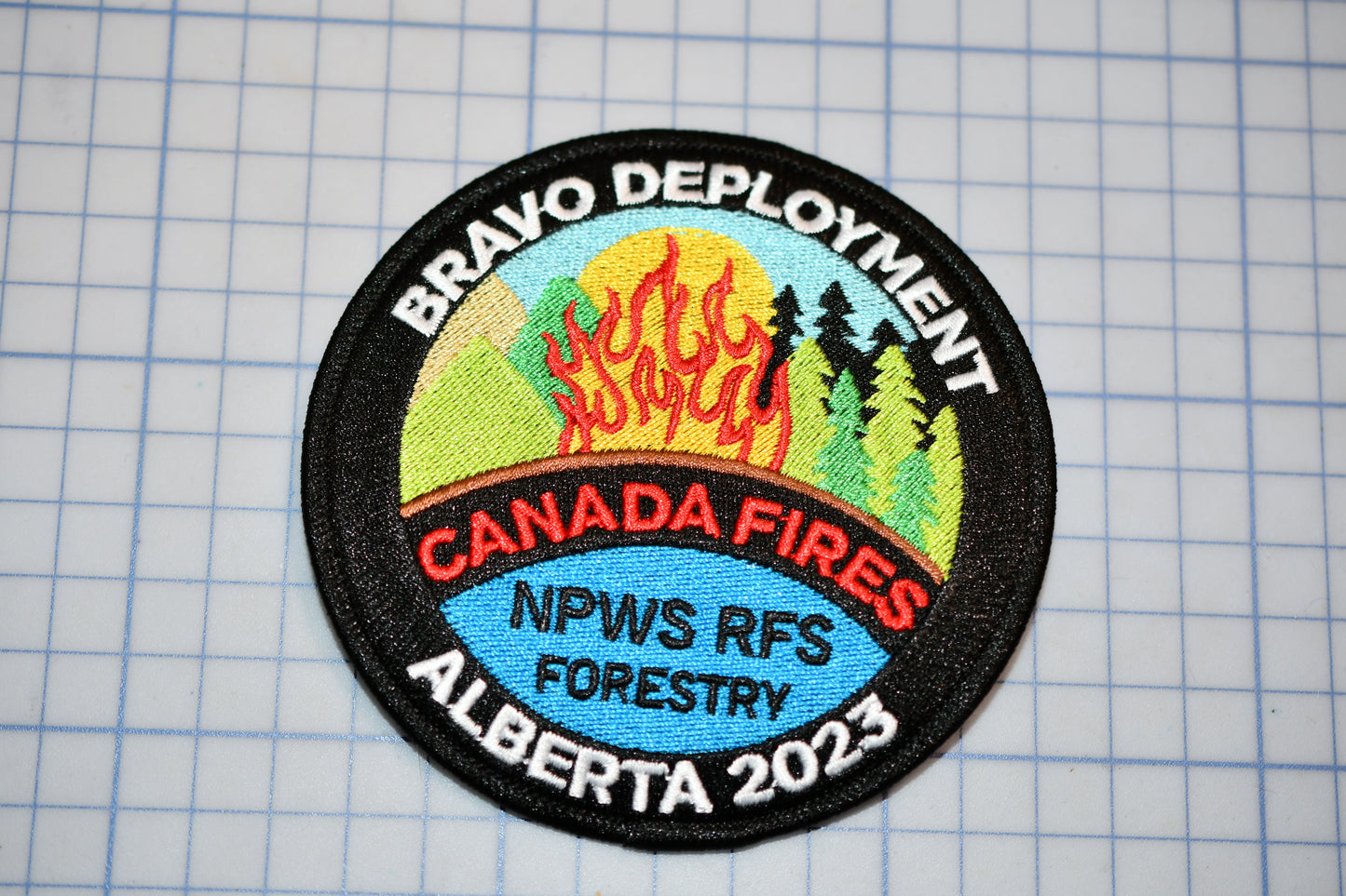 A badge with the text "BRAVO DEPLOYMENT" at the top, "CANADA FIRES" in the center, and "NPWS RFS FORESTYLE" at the bottom. The badge is placed on a grid background.