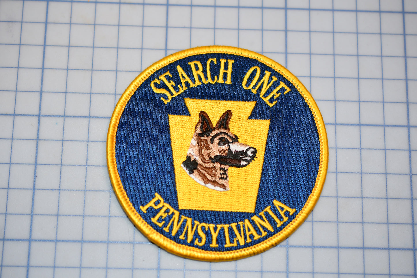 a circular patch with a dog's head and the text "SEARCH ONE PENNSYLVANIA" on it, placed on a grid background.
