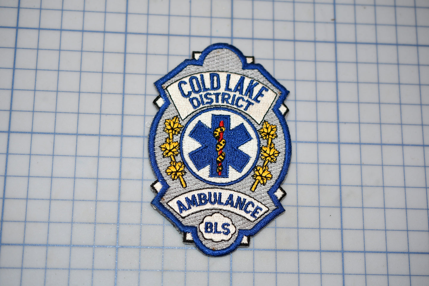 a badge or patch with the text "COLD LAKE DISTRICT AMBULANCE" and a medical symbol, indicating that it is a badge or patch from the Cold Lake District Ambulance.