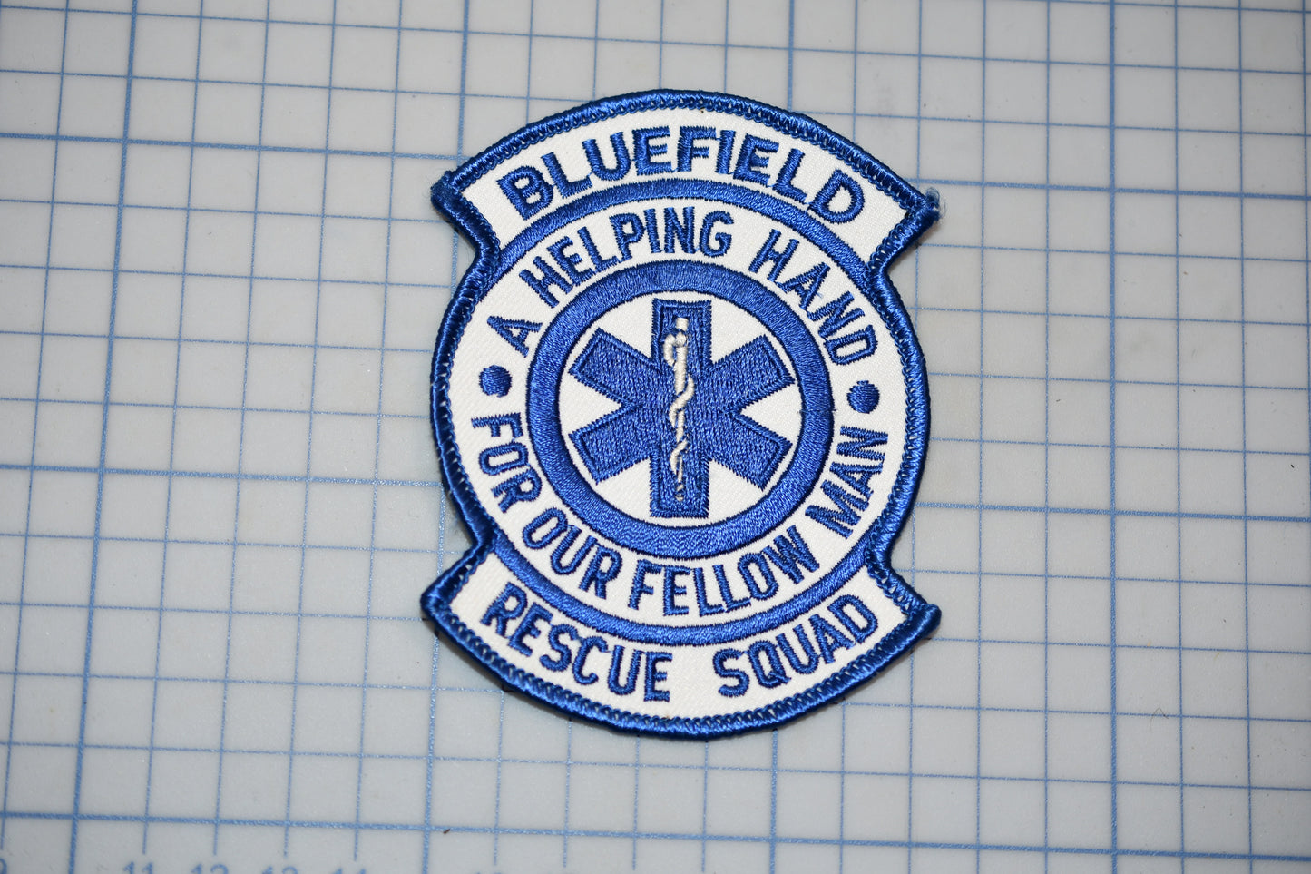 A blue and white patch with the text "BLUEFIELD A HELPING HAND FOR OUR FELLOW MAN RESCUE SQUAD" and a medical symbol, indicating that it is a patch representing the Bluefield Rescue Squad.