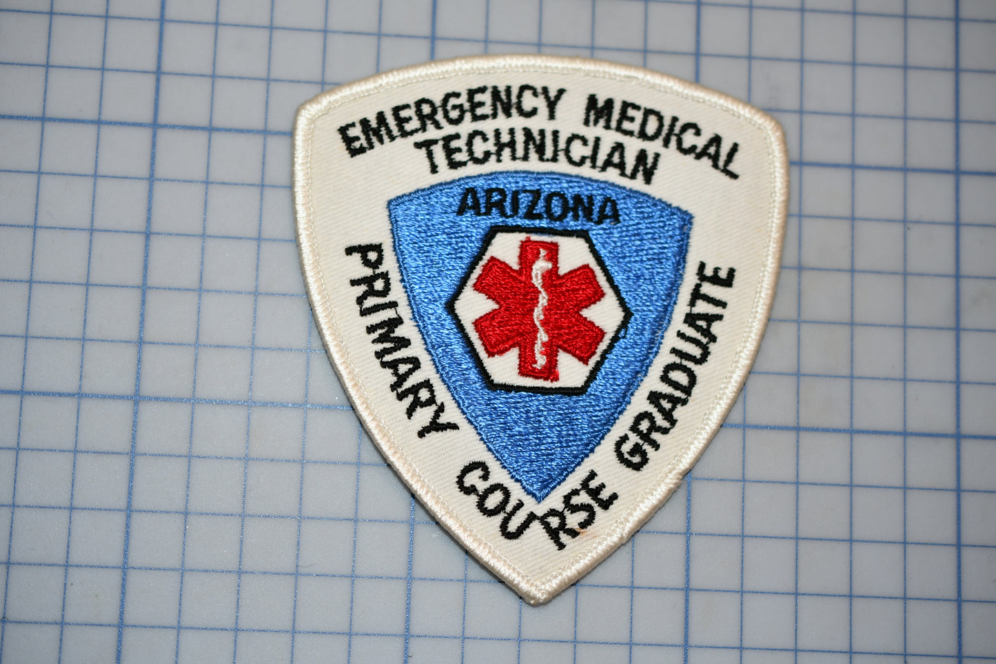 a patch with the text "EMERGENCY MEDICAL TECHNICIAN ARIZONA PRIMARY GRADUATE" and a red cross symbol, indicating that it is a patch for an emergency medical technician.