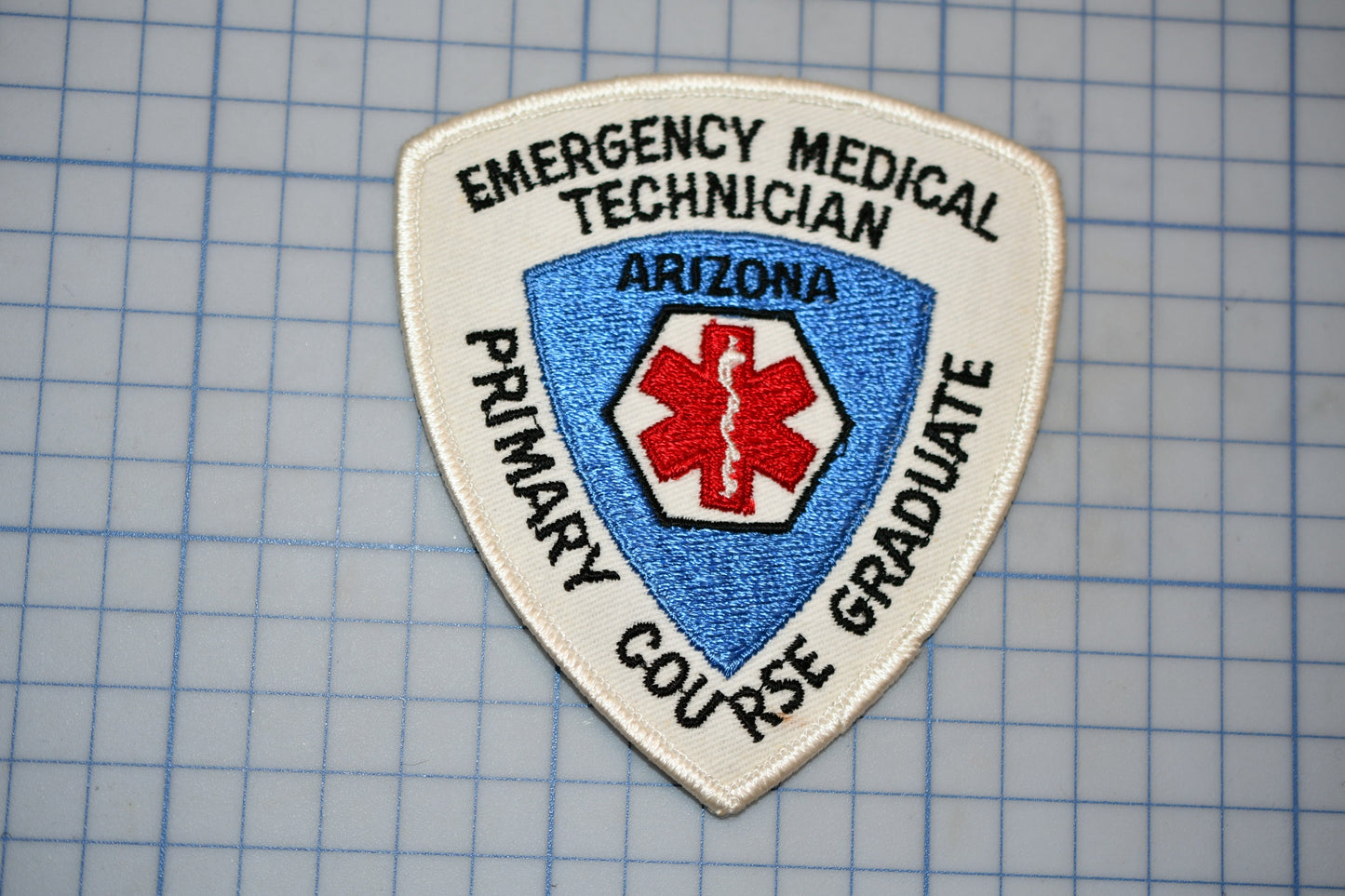 a patch with the text "EMERGENCY MEDICAL TECHNICIAN ARIZONA PRIMARY GRADUATE" and a red cross symbol, indicating that it is a patch for an emergency medical technician.