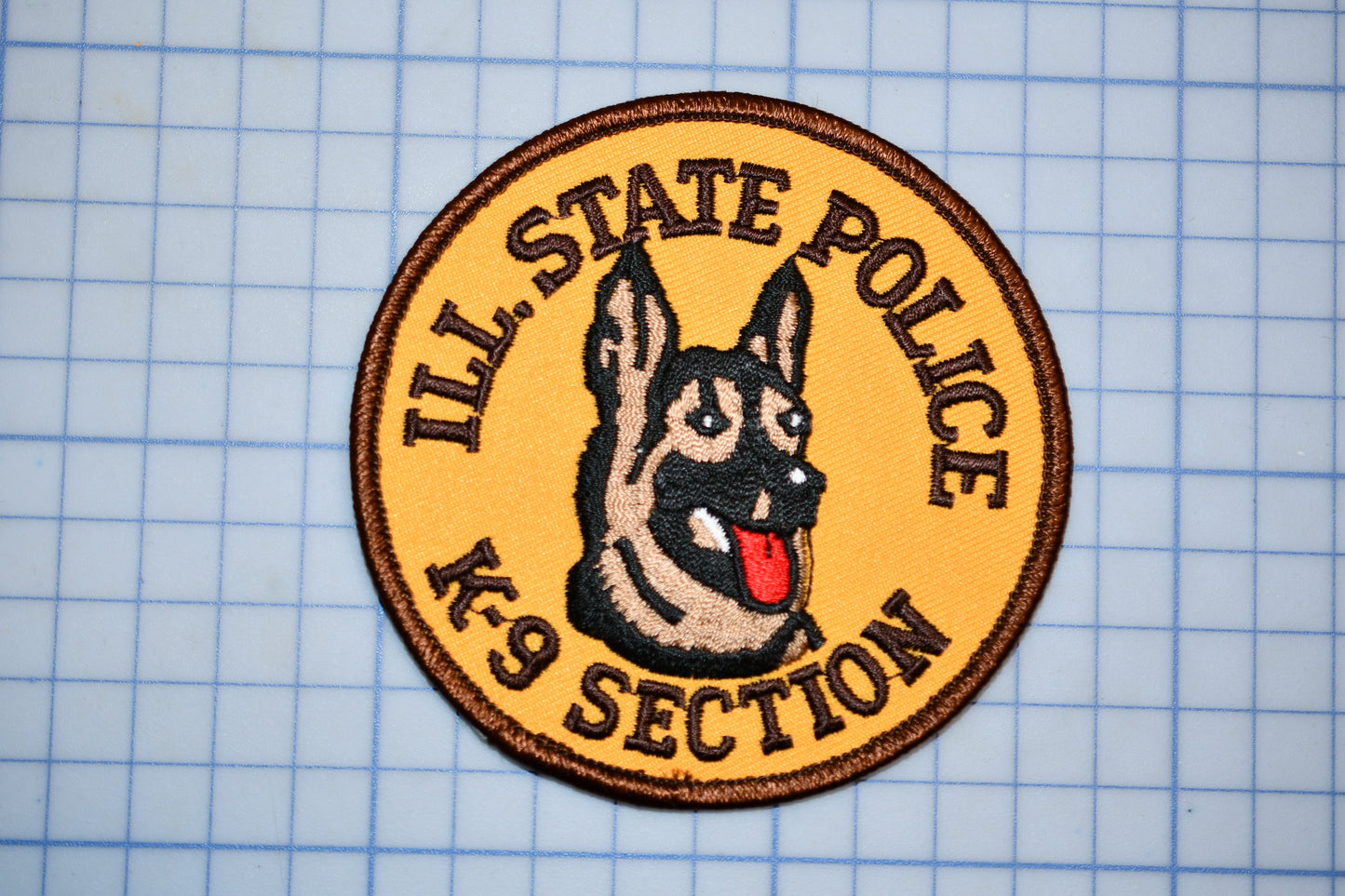 A circular patch with a dog's head and tongue, surrounded by text that reads "ILL. STATE POLICE K-9 SECTION".