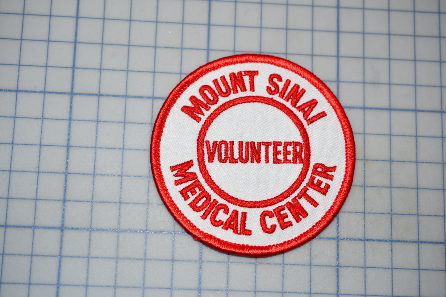 A red and white patch with the text "MOUNT SINAI VOLUNTEER MEDICAL CENTER" printed on it, placed on a grid background.