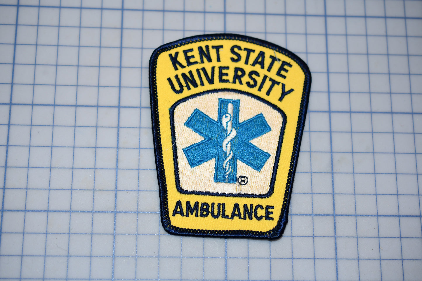 a yellow and blue patch with the text "KENT STATE UNIVERSITY AMBULANCE" and a medical symbol, indicating that it is a patch representing the Kent State University Ambulance Service.