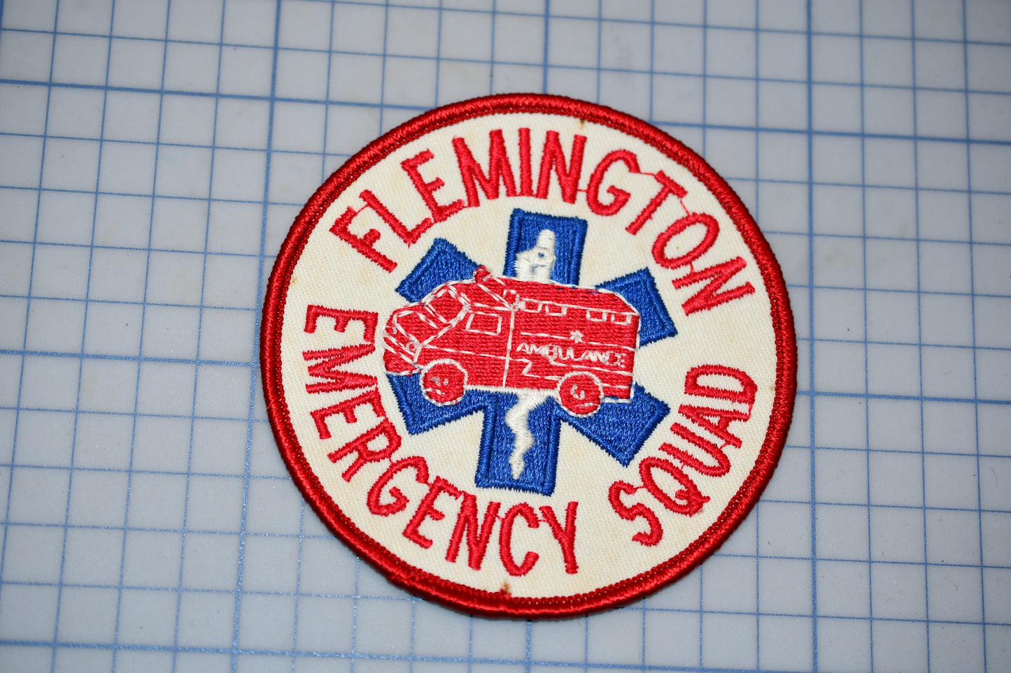 A patch with the text "FLEMINGTON EMERGENCY SQUAD" and an image of a red and blue ambulance, indicating that it is a patch representing the emergency medical services of the town of Flemington.