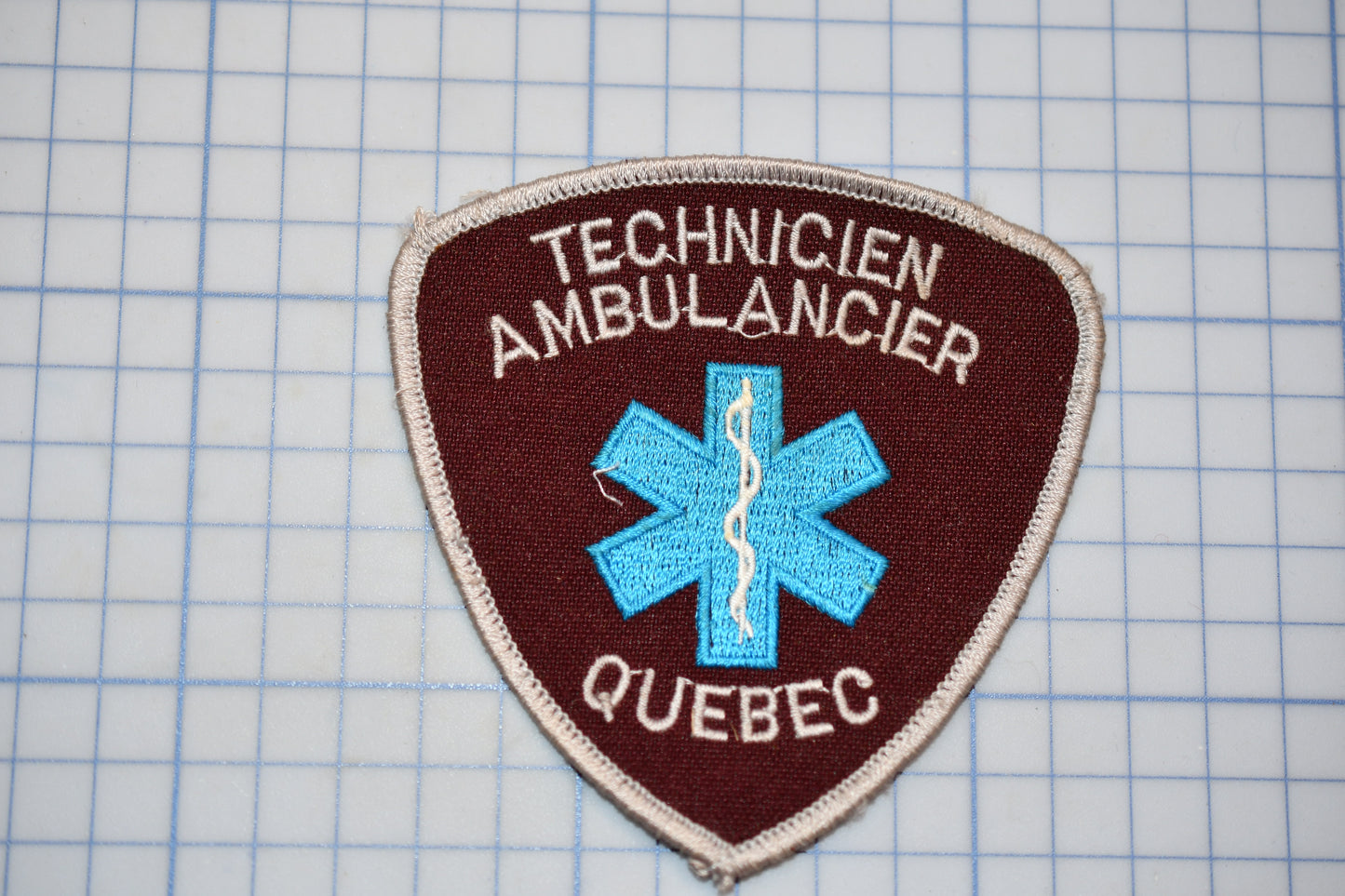 a red and blue patch with the text "TECHNICIEN AMBULANCIER QUEBEC" and a blue medical symbol, indicating that it is a patch for a technician ambulance from Quebec.