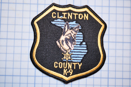 A black and gold badge with a dog and the text "CLINTON COUNTY K-9" on it.