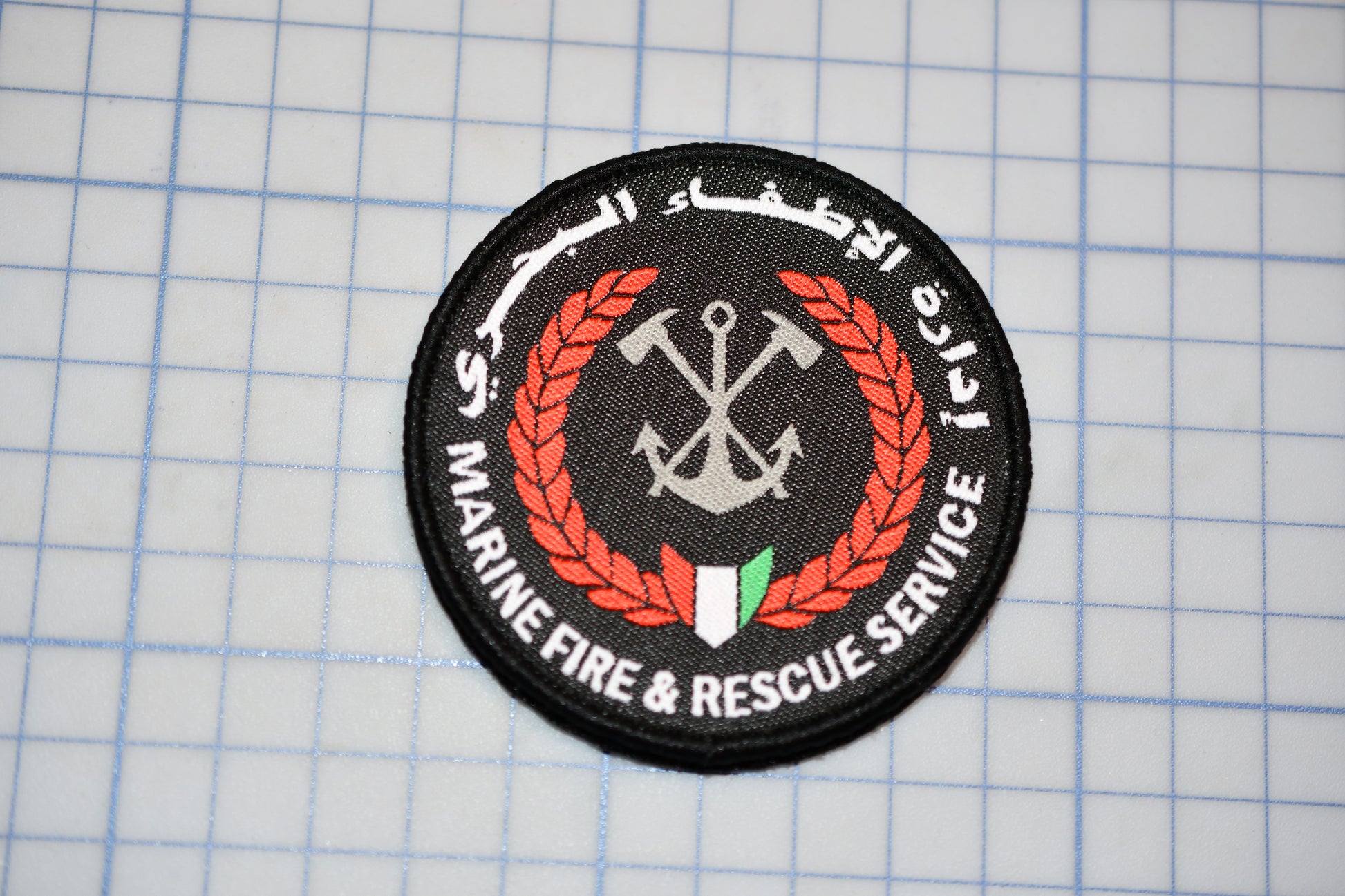 a circular patch with a black background, a red laurel wreath, and a white anchor symbol. The text around the patch reads "Marine Fire & Rescue Service".