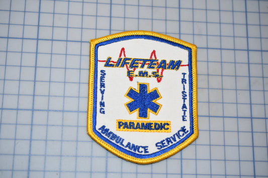 a patch with the word "LIFETIME" written on it, along with a medical symbol. The patch is part of a larger collection of patches, as indicated by the text "LIFETIME EMERGENCY SERVICE" and "PARAMEDIC AMBULANCE SERVICE".