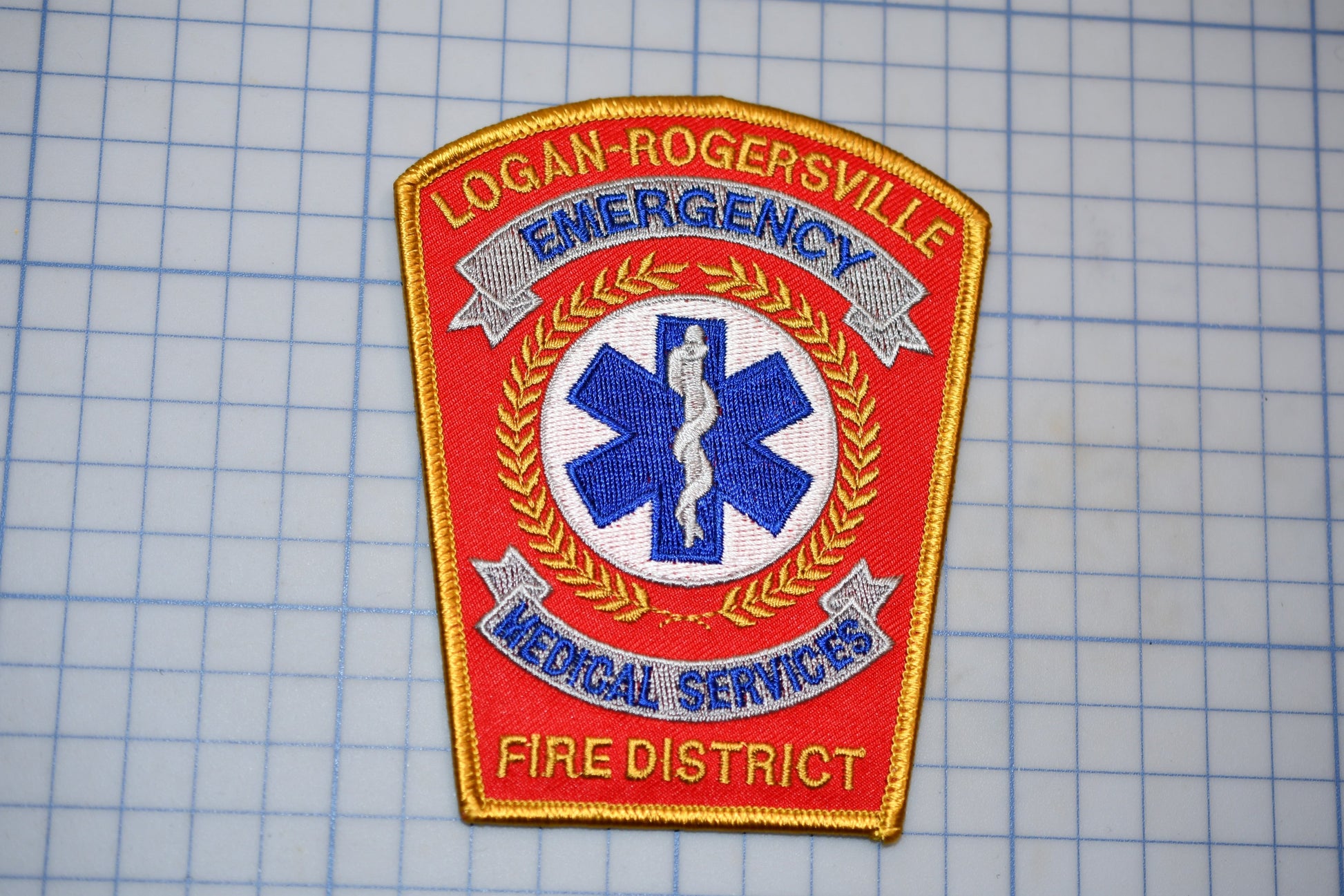 a red and gold emergency services patch with a blue and white medical cross symbol, indicating that it belongs to the Logan-Rogersville Fire District.