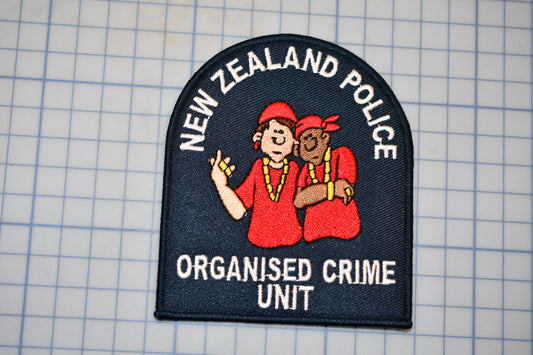 a New Zealand Police Organised Crime Unit patch, which is a black and white patch with a cartoon image of two people, one wearing a red shirt and the other wearing a red hat.