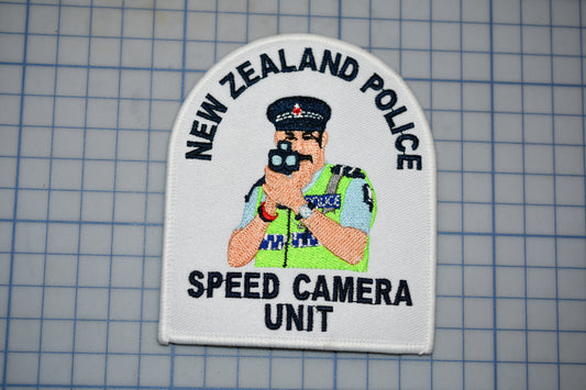 a white patch with an embroidered illustration of a police officer holding a speed camera. The patch also displays the text "NEW ZEALAND POLICE SPEED CAMERA UNIT".