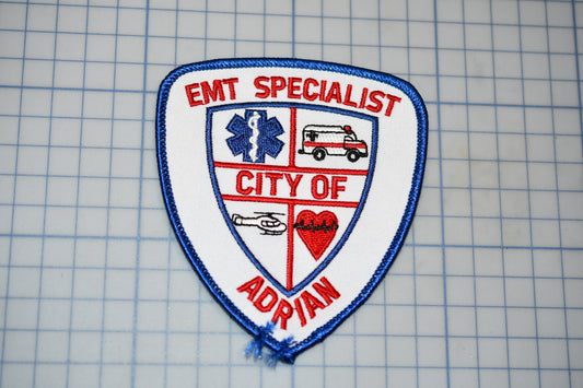 a badge or patch with the text "EMT SPECIALIST CITY OF ADRIAN" and various medical and emergency-related symbols, such as a cross, ambulance, and heart.