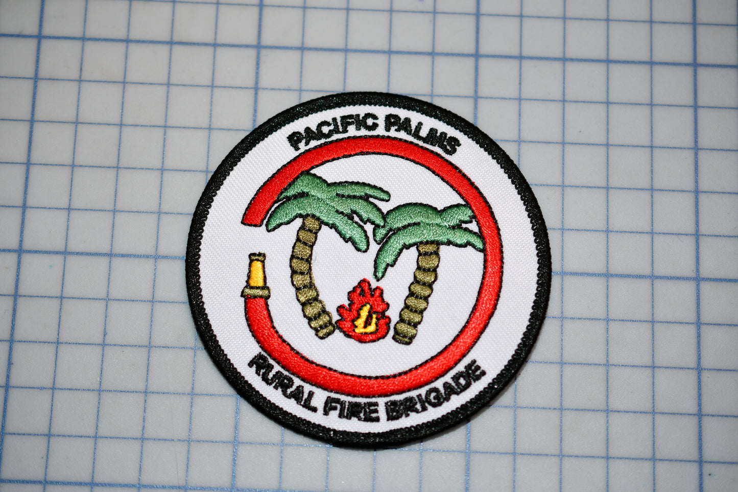 a circular patch with a red border, depicting a firefighting emblem with palm trees and a fire. The patch is labeled "Pacific Palms Rural Fire Brigade".