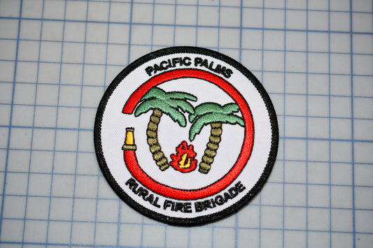 a circular patch with a red border, depicting a firefighting emblem with palm trees and a fire. The patch is labeled "Pacific Palms Rural Fire Brigade".