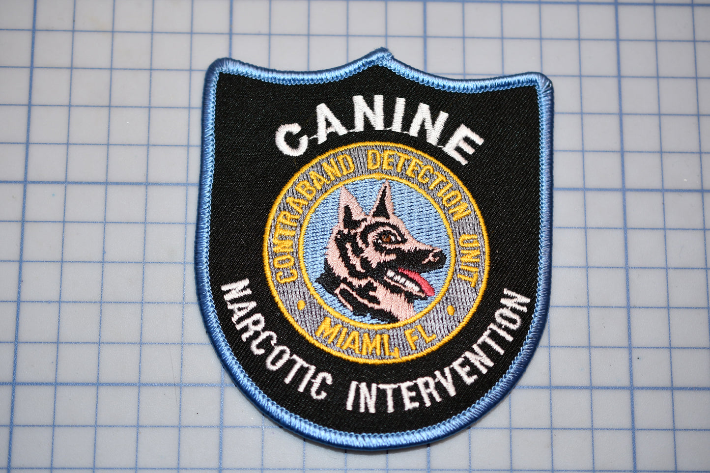 A black and blue patch with the text "CANINE DETECTION UNIT" and "NARCOTIC INTERVENTION" printed on it, along with a graphic of a dog's head.