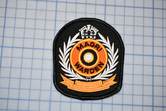 a black and orange patch with the text "MAORI WARDEN" and a crown-like emblem, placed on a grid background.