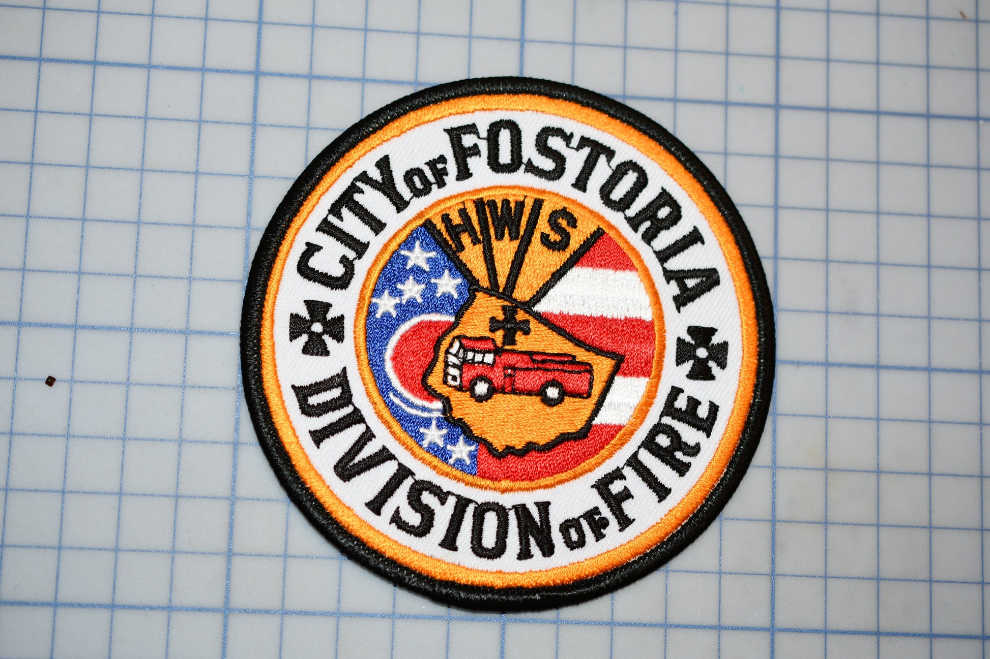 a circular patch with a design that includes a fire truck, stars, and the text "City of Fostoria Division of Fire".