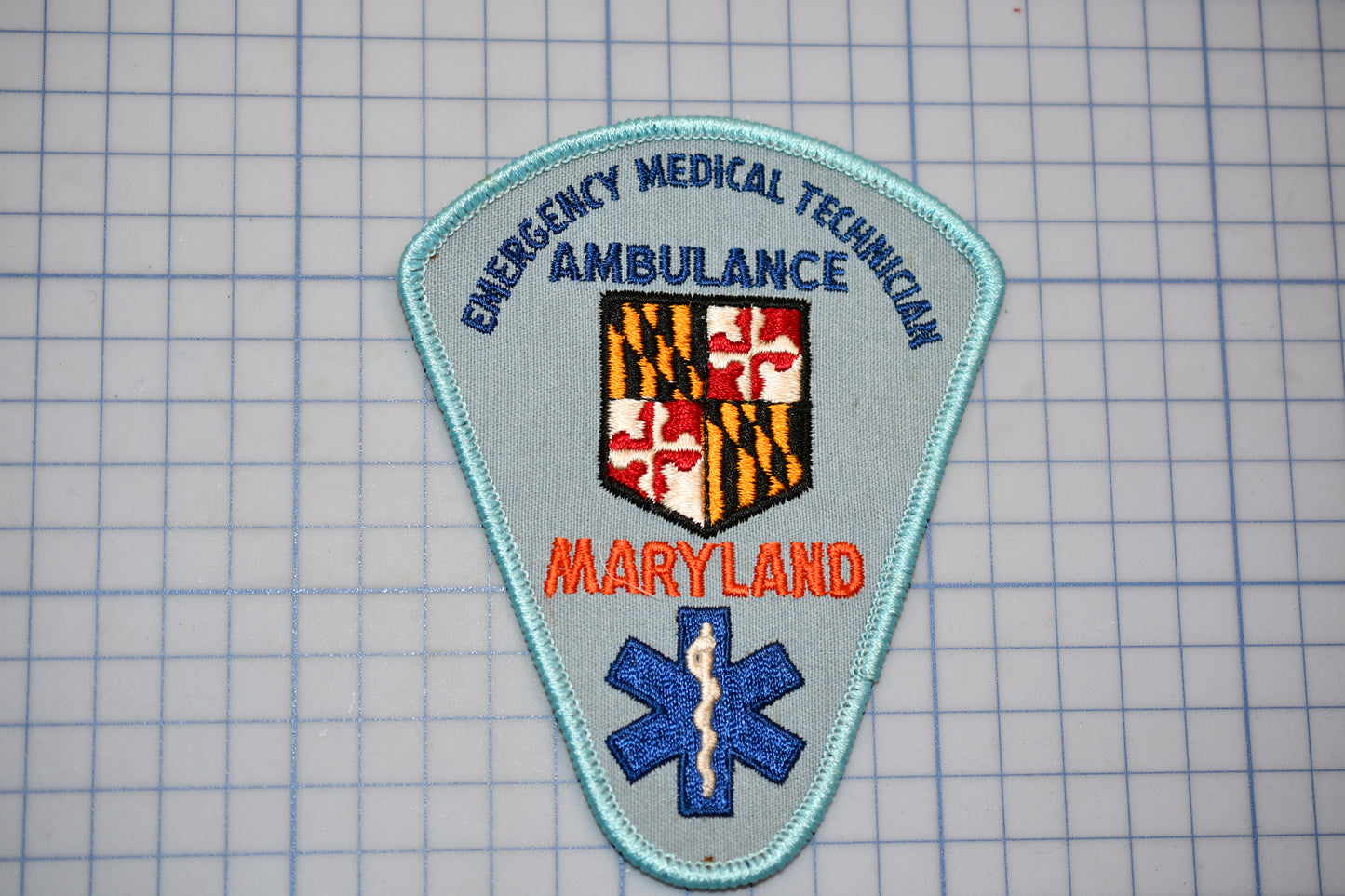 a patch with the text "EMERGENCY MEDICAL TECHNICIAN AMBULANCE" and "MARYLAND" printed on it, along with a shield-shaped emblem and a blue medical cross symbol.