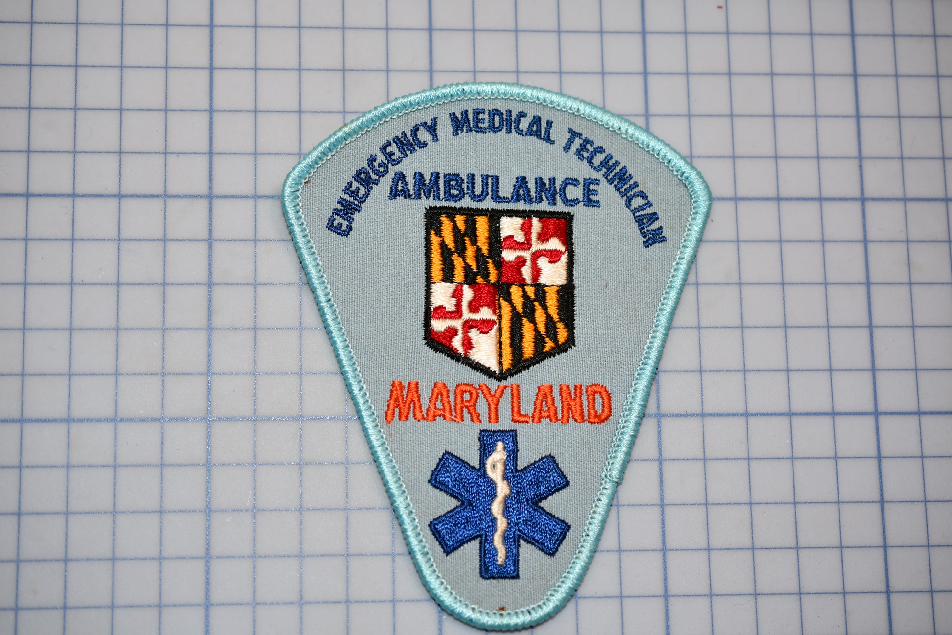 a patch with the text "EMERGENCY MEDICAL TECHNICIAN AMBULANCE" and "MARYLAND" printed on it, along with a shield-shaped emblem and a blue medical cross symbol.