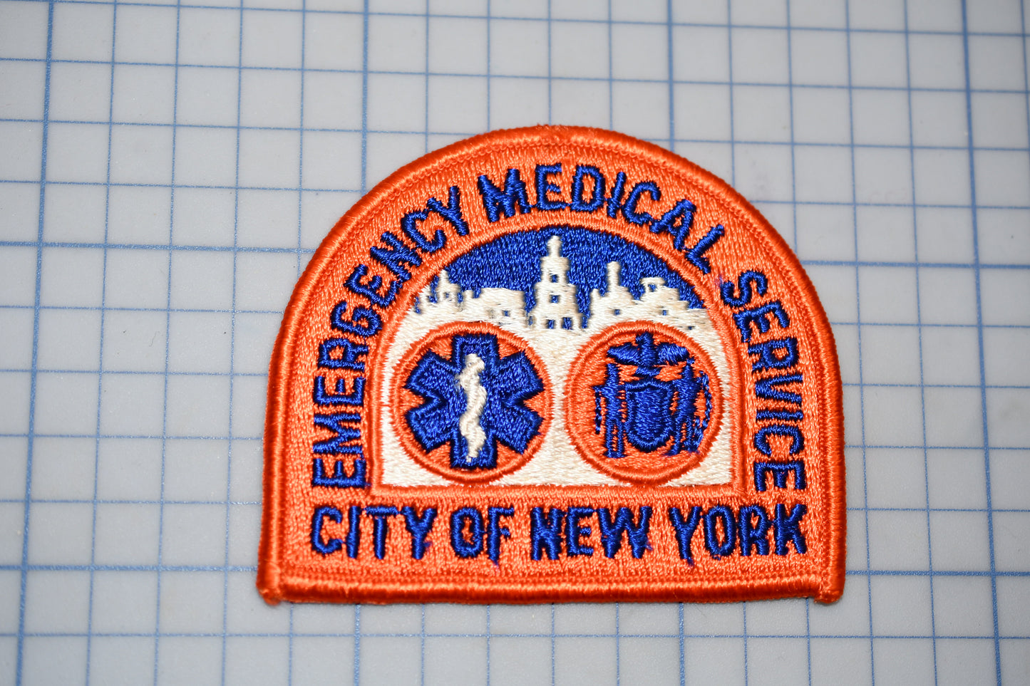 a patch with the text "EMERGENCY MEDICAL SERVICE CITY OF NEW YORK" and two medical symbols, one of which is a cross and the other a stethoscope.