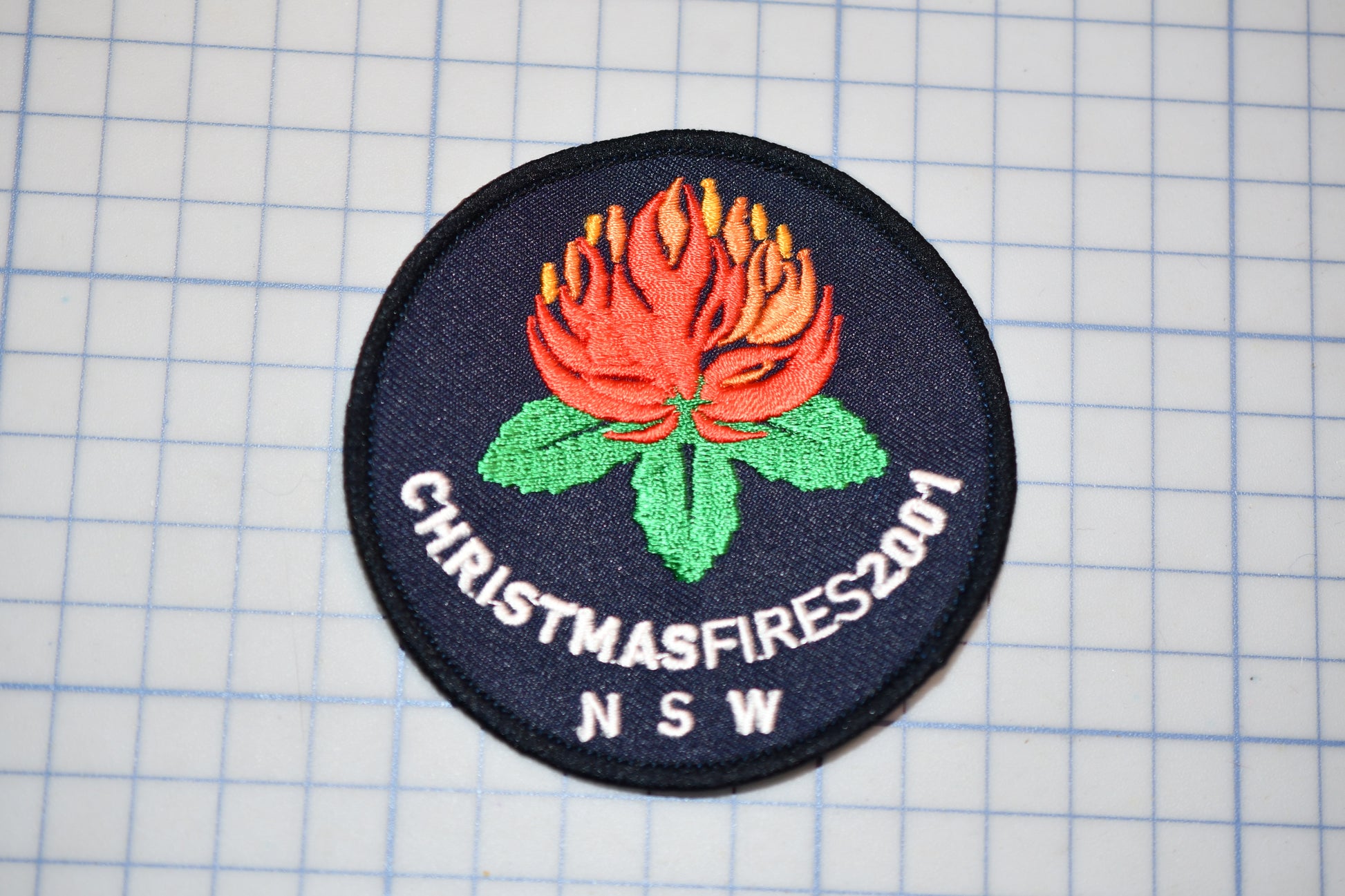 A circular patch with a red flower and green leaves, along with the text "CHRISTMASFIRES2001 N SW" printed on it.