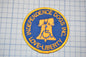 a circular patch with a blue background and gold border, displaying the text "INDEPENDENCE DOGS INC. LOVE LIBERTY" along with a symbol of a bell tower.