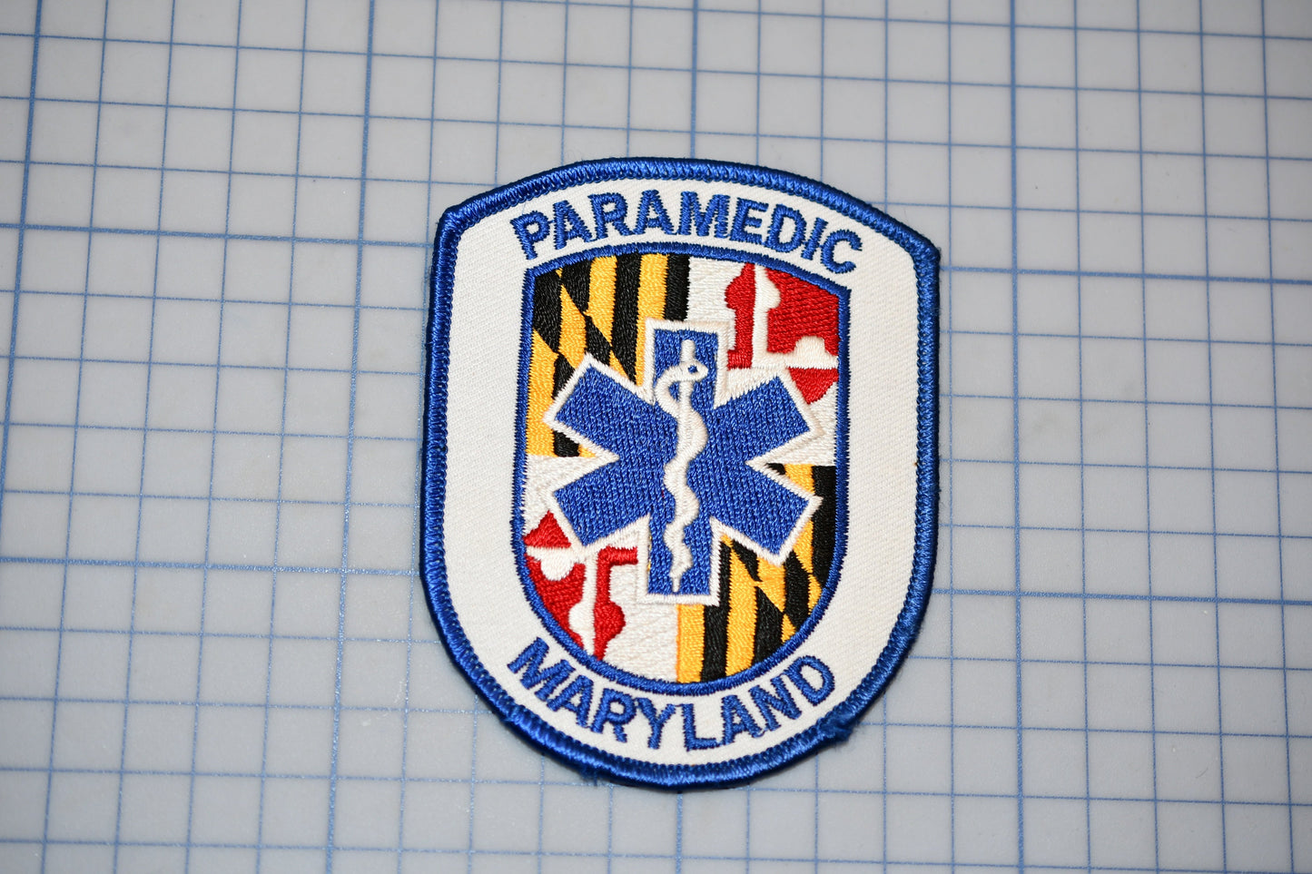 a Paramedic Maryland patch, which is a blue and white shield-shaped emblem with a medical cross and the text "PARAMEDIC MARYLAND" printed on it.