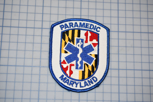 a Paramedic Maryland patch, which is a blue and white shield-shaped emblem with a medical cross and the text "PARAMEDIC MARYLAND" printed on it.
