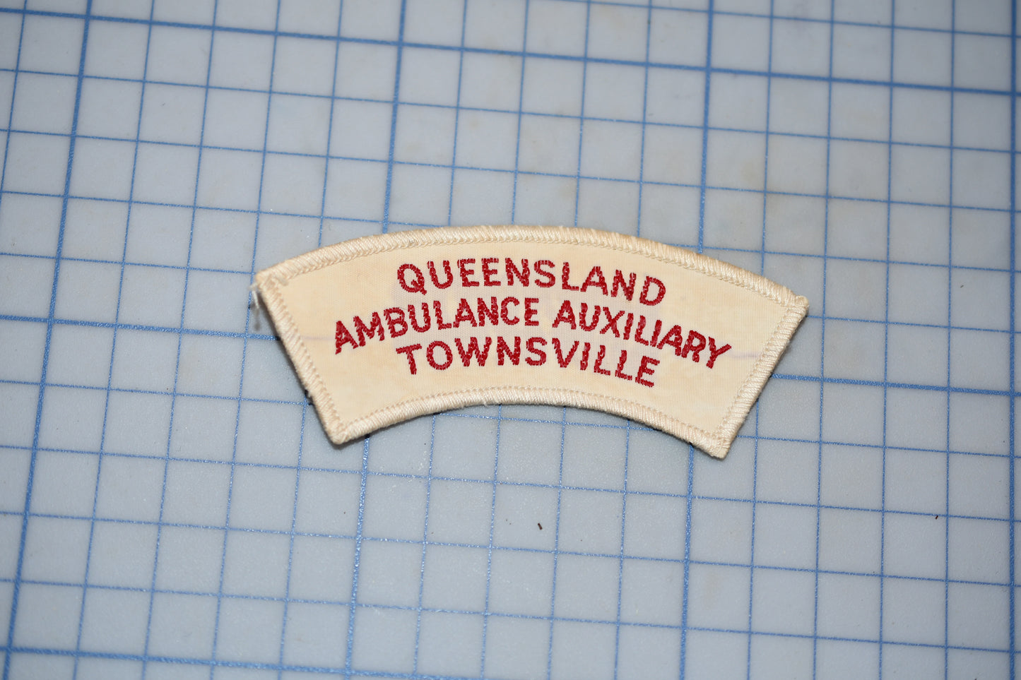 a patch with the text "Queensland Ambulance Auxiliary Townsville" printed on it, placed on a grid background.