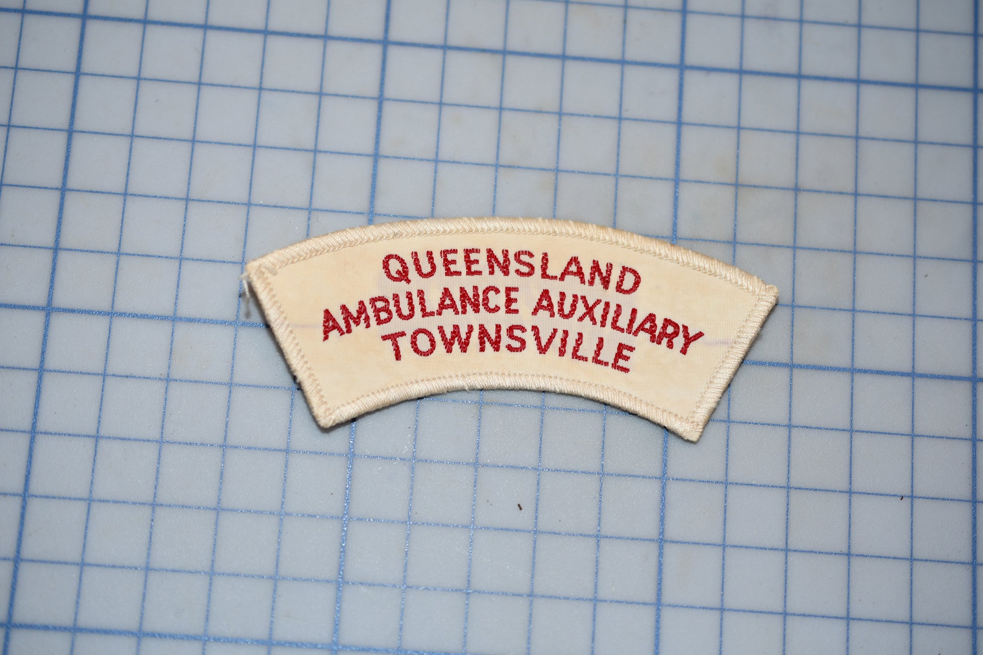 a patch with the text "Queensland Ambulance Auxiliary Townsville" printed on it, placed on a grid background.