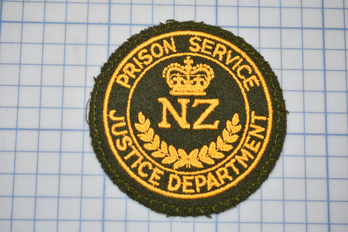 a circular badge with the text "PRISON SERVICE" and "NZ JUSTICE DEPARTMENT" embroidered on it, along with a crown symbol.
