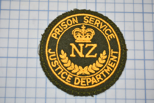 a circular badge with the text "PRISON SERVICE" and "NZ JUSTICE DEPARTMENT" embroidered on it, along with a crown symbol.