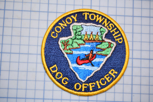 a circular patch with a blue border and yellow border, depicting a dog in a red boat on a river. The patch has the text "CONOY TOWNSHIP" and "DOG OFFICER" written around the edge.