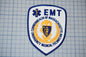 a badge or patch with the text "EMT" and a shield-shaped emblem, surrounded by a blue and gold border.