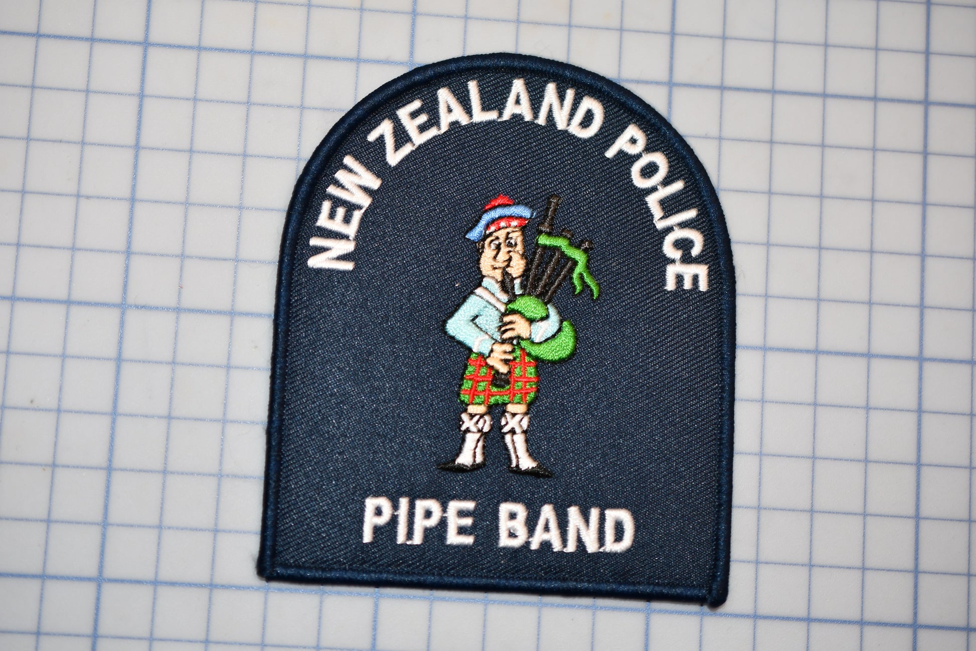 a patch with a cartoon character playing a musical instrument, specifically a pipe. The patch is labeled "New Zealand Police Pipe Band".