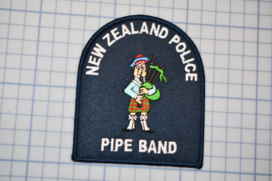a patch with a cartoon character playing a musical instrument, specifically a pipe. The patch is labeled "New Zealand Police Pipe Band".