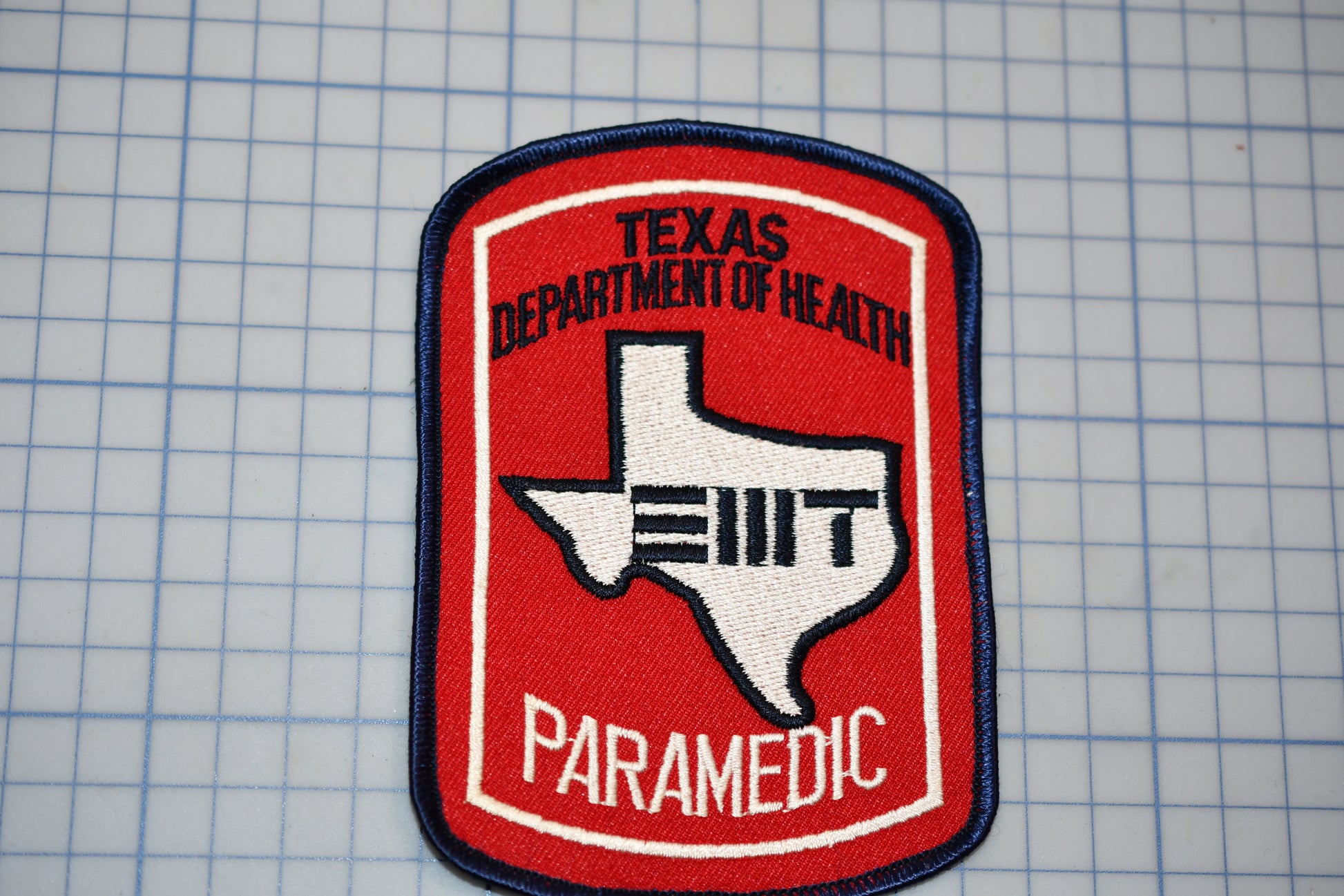 A red and white Texas Department of Health Paramedic (TDI) patch is displayed against a grid background.