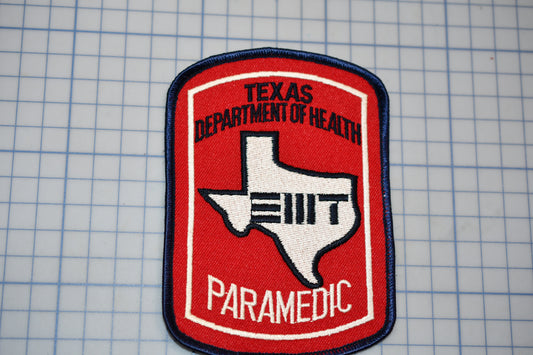 A red and white Texas Department of Health Paramedic (TDI) patch is displayed against a grid background.