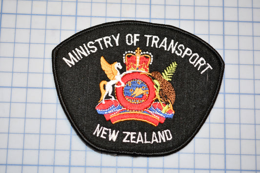 a black patch with the text "Ministry of Transport New Zealand" and a coat of arms, all set against a grid background.