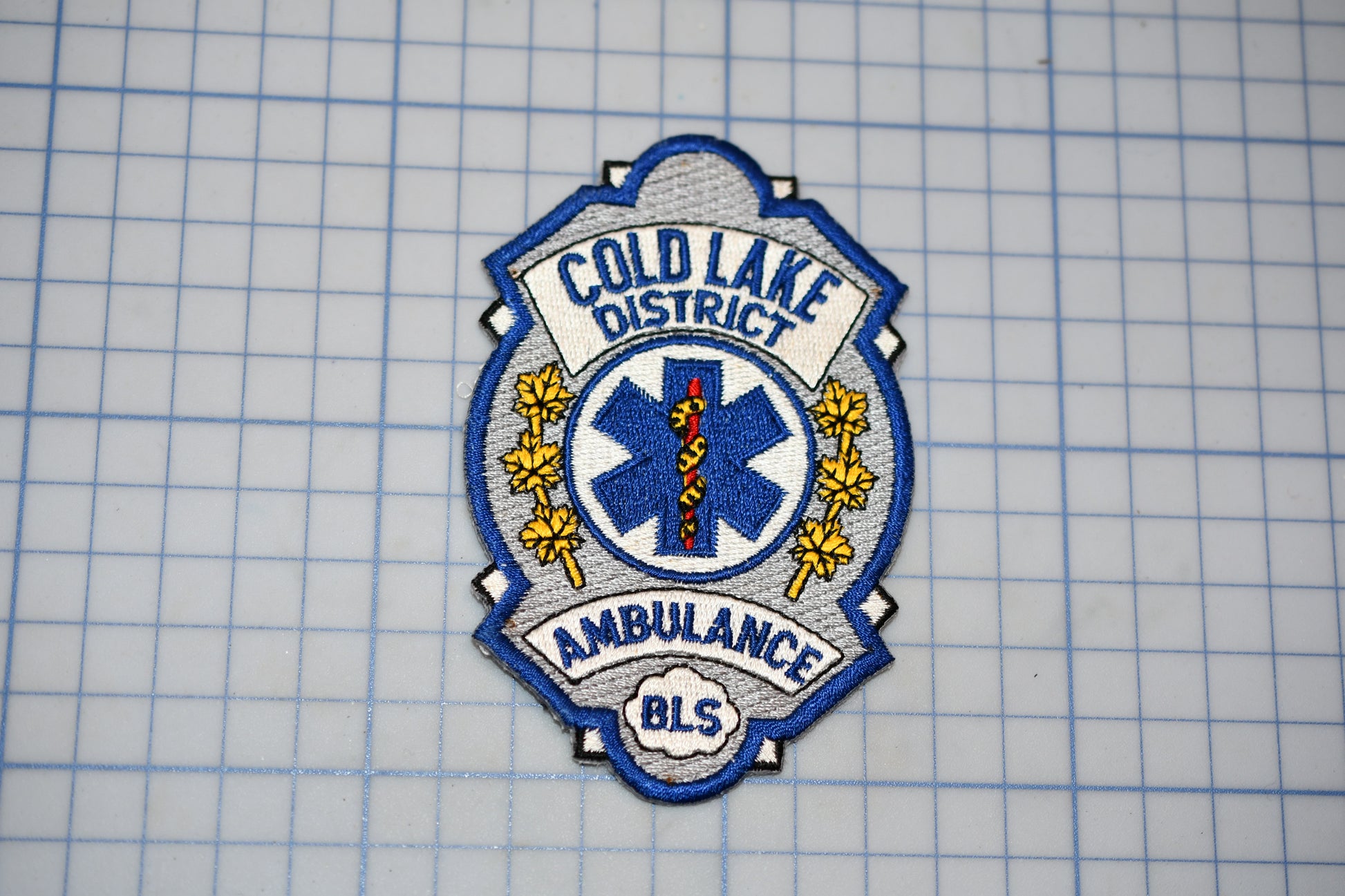 a badge or patch with the text "COLD LAKE DISTRICT AMBULANCE" and a medical symbol, indicating that it is a badge or patch from the Cold Lake District Ambulance.