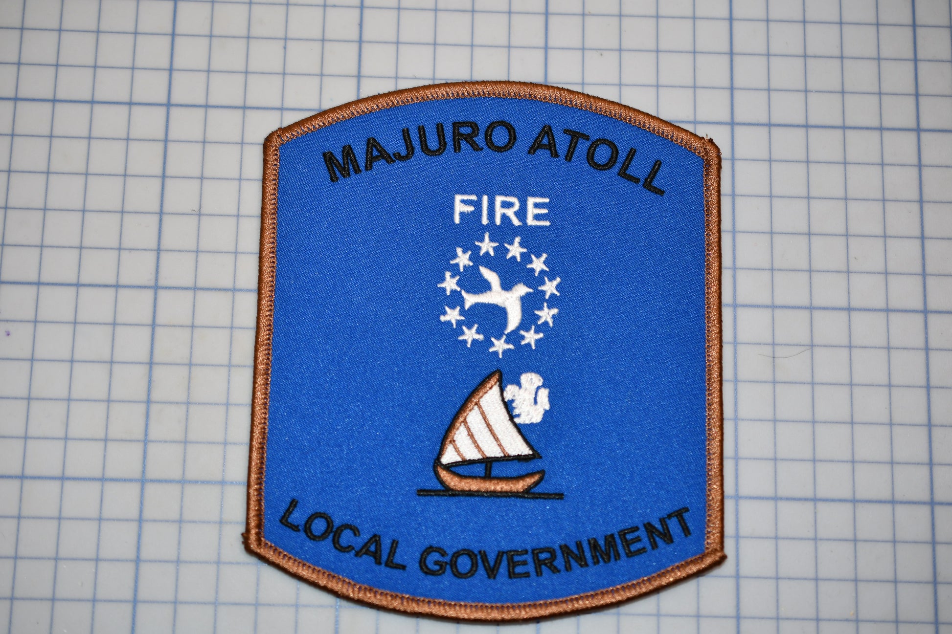 a blue and brown patch with text and a sailboat symbol, representing the Majuro Atoll Fire Local Government.