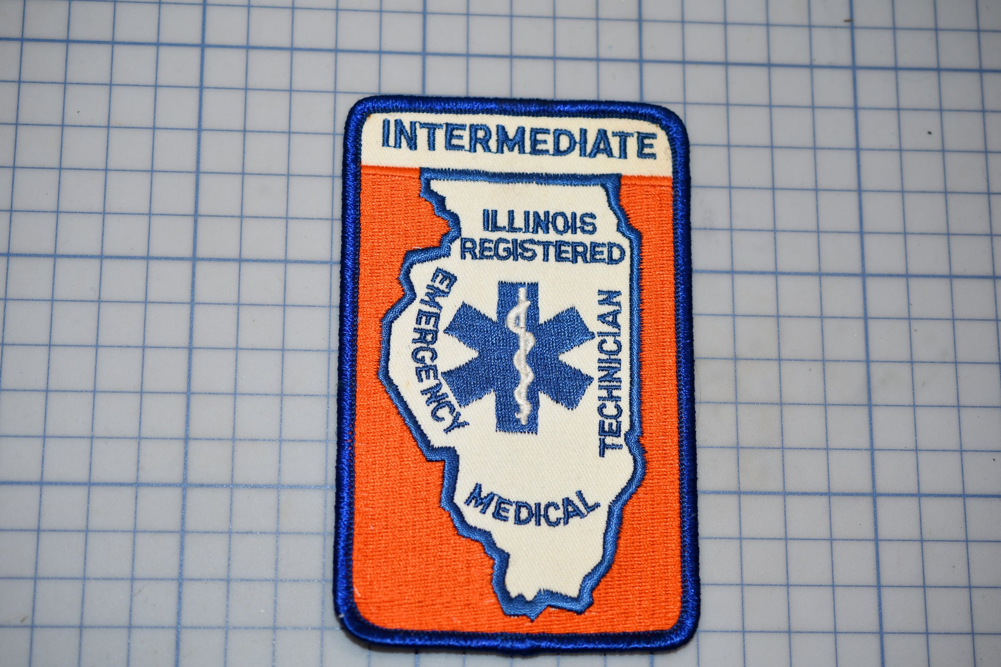 a patch with the word "INTERMEDIATE" and the word "ILLINOIS" written on it, along with the word "EMERGENCY" and "TECHNICAL" written below. The patch is set against a blue grid background.